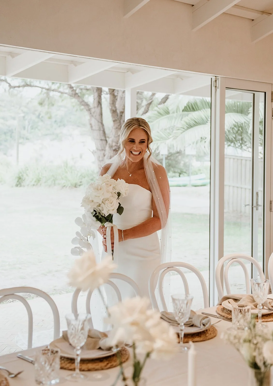 A bride stands indoors holding a bouquet of white flowers, smiling, in front of a large window with a garden view.