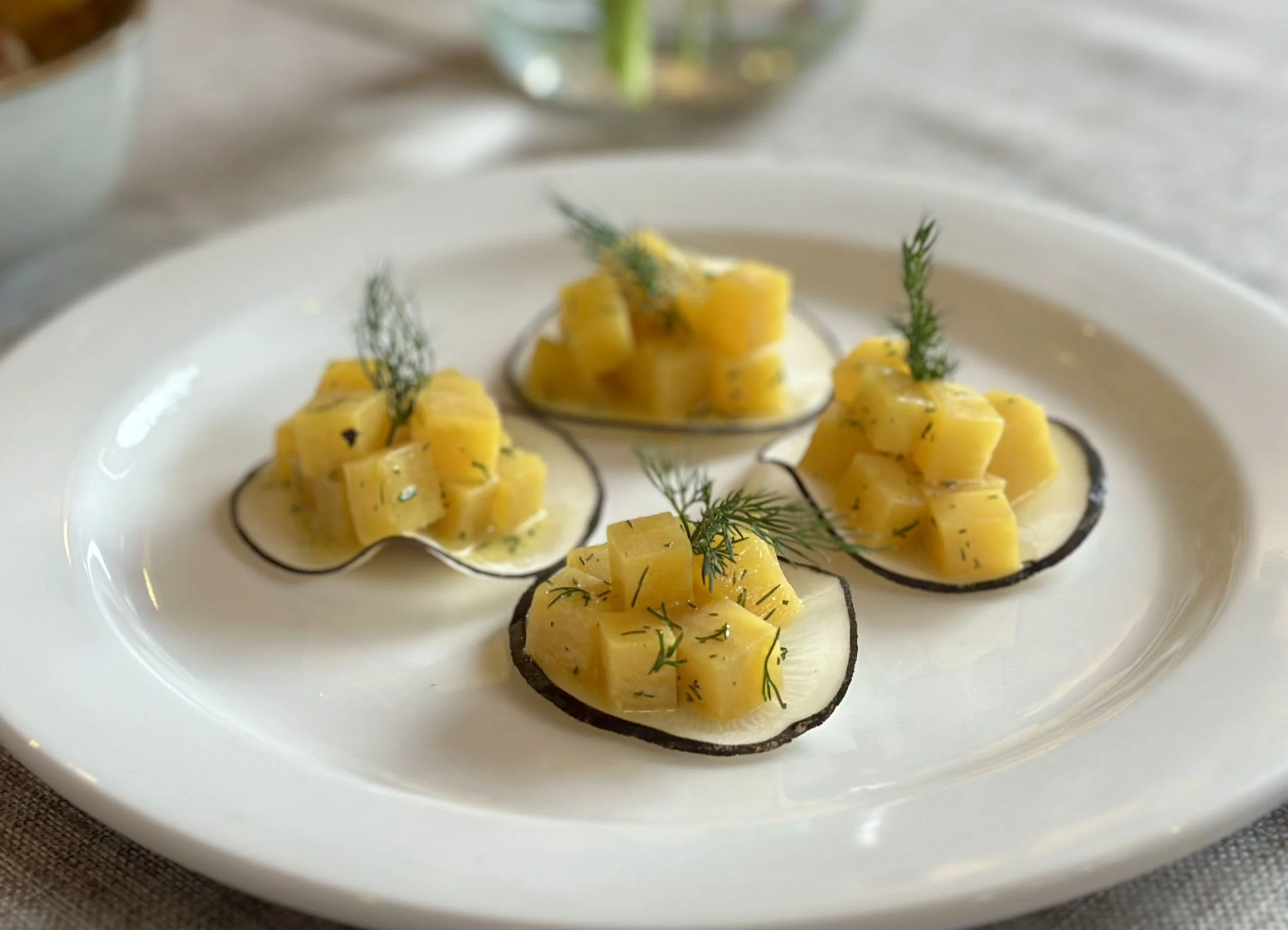 A white plate with four tartlet shells filled with a yellow filling, garnished with herbs, and topped with small diced yellow fruit or vegetable.