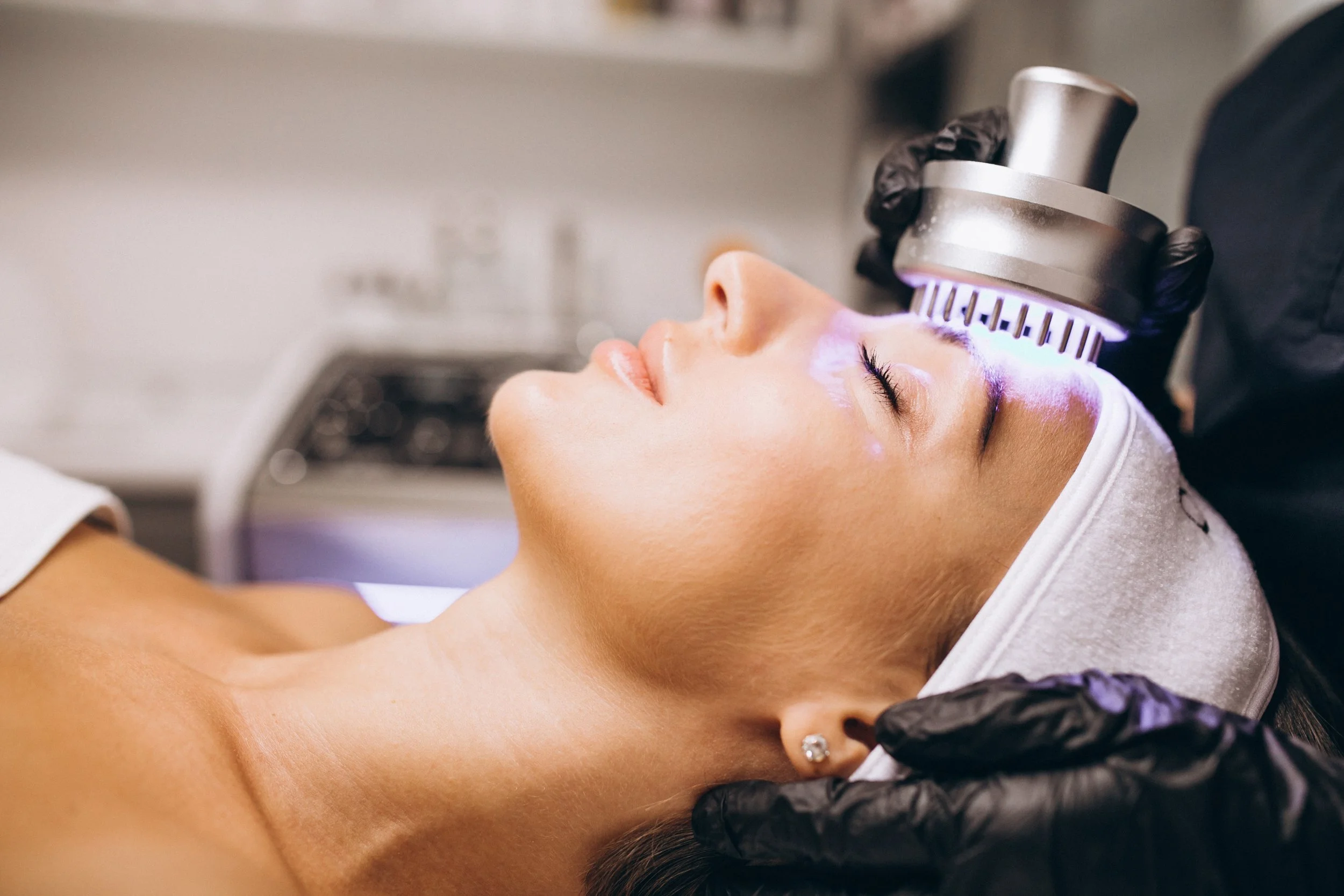 A woman lying down with her eyes closed during a facial treatment, receiving LED light therapy on her face from a technician wearing black gloves.