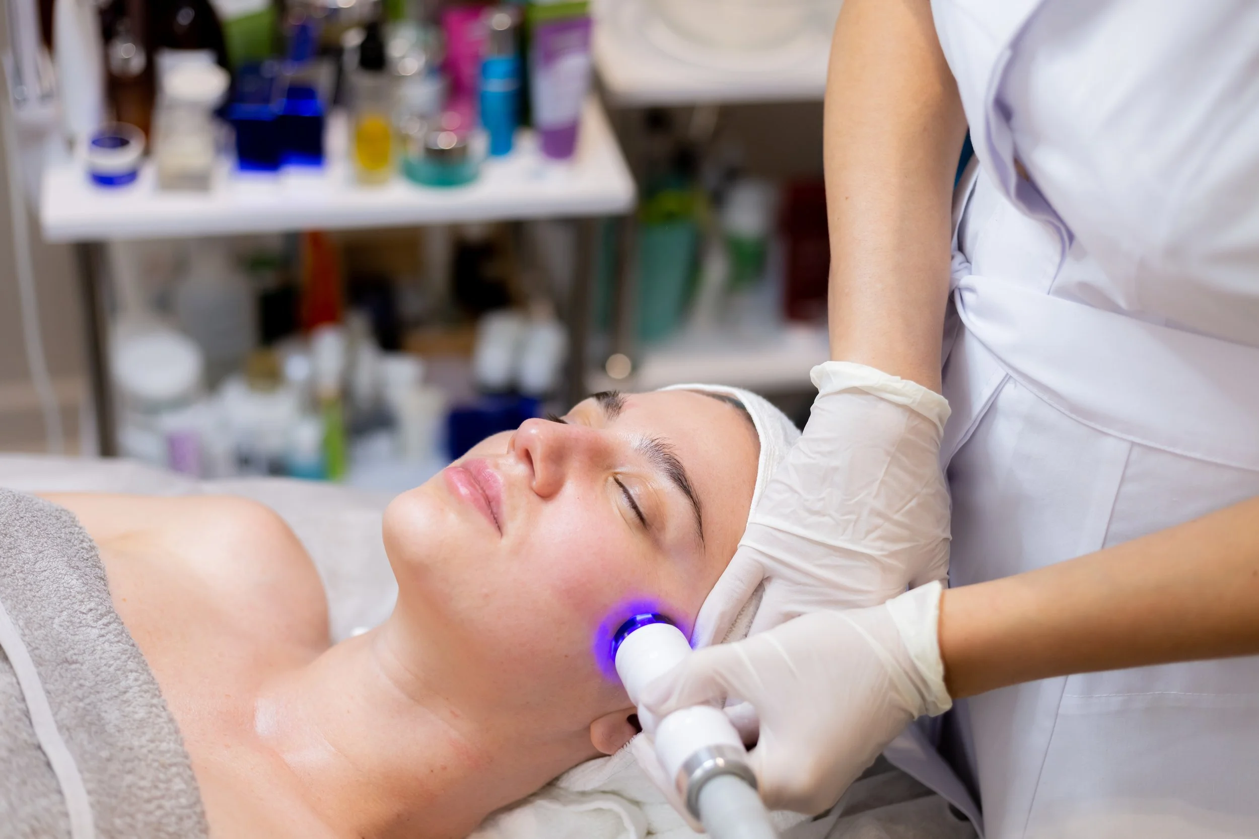 A woman receiving a facial treatment in a spa or clinic setting, lying on a treatment bed with a towel under her head, while a professional in white scrubs applies a device to her face.