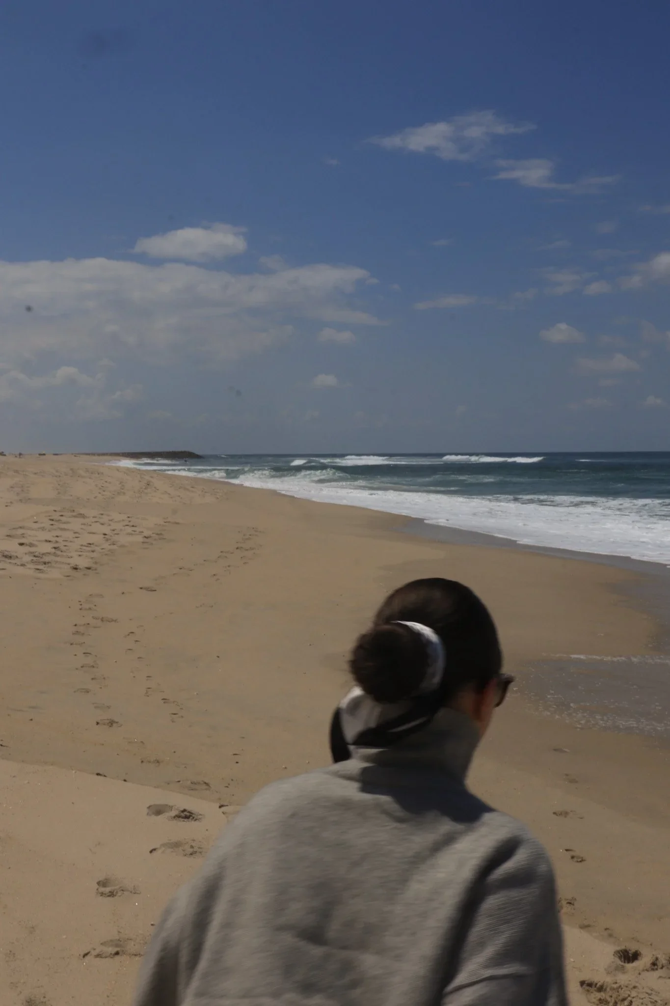 Une personne avec une queue de cheval portée dans un foulard noir, vue de dos, marchant sur une plage avec du sable fin, face à l'océan sous un ciel partiellement nuageux.