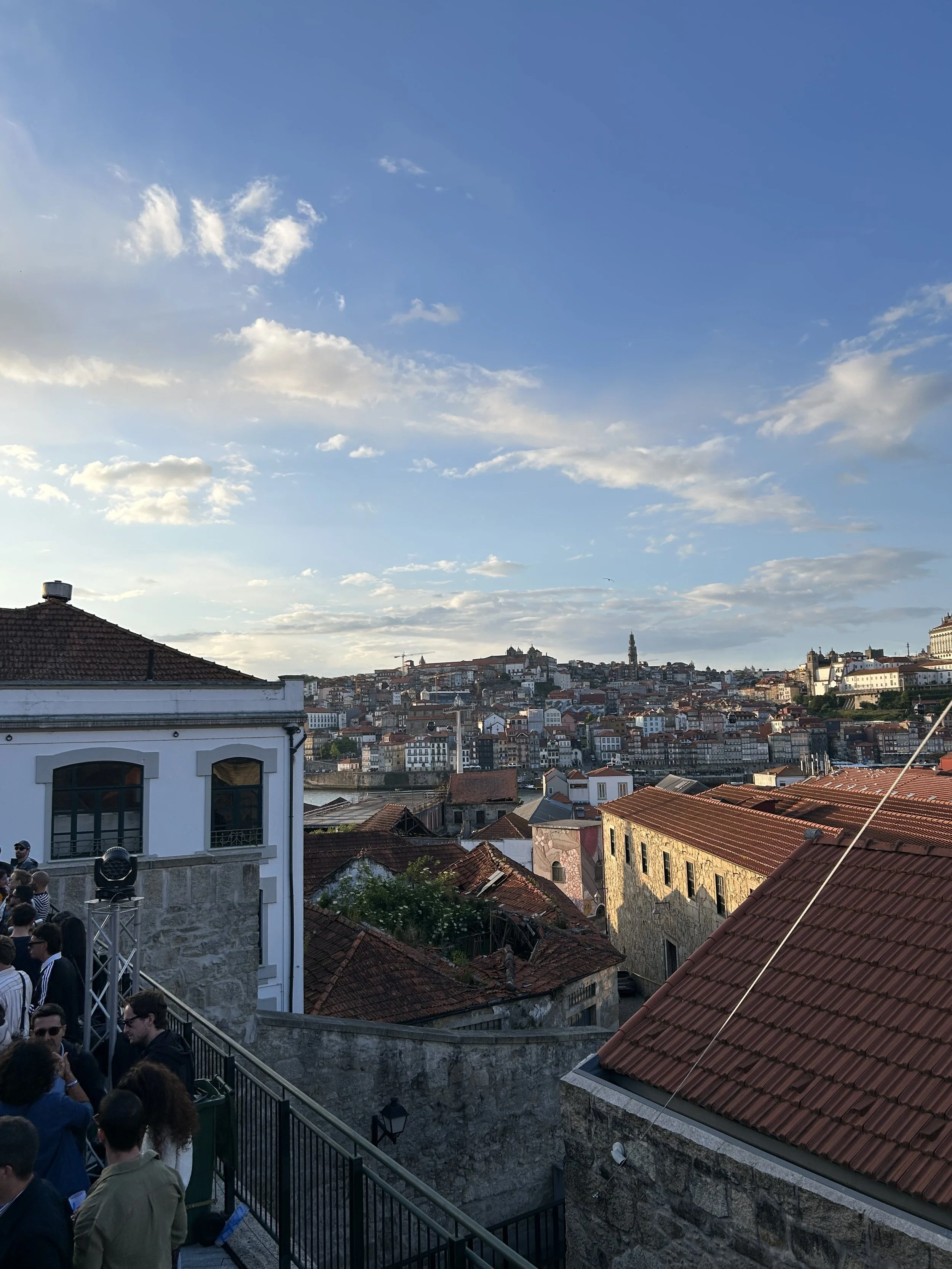 Vue sur une ville avec des bâtiments en tuiles rouges, une partie de la ville en contrebas et d'autres bâtiments en hauteur au loin, sous un ciel bleu avec quelques nuages.