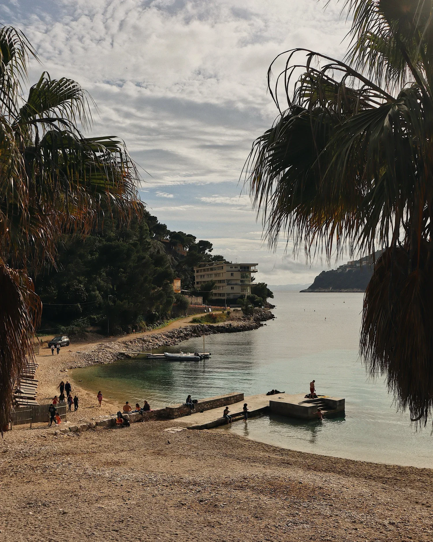 Plage de sable avec des personnes, palmier, bâtiments en hauteur sur une côte rocheuse, mer calme, bateau, ciel nuageux.