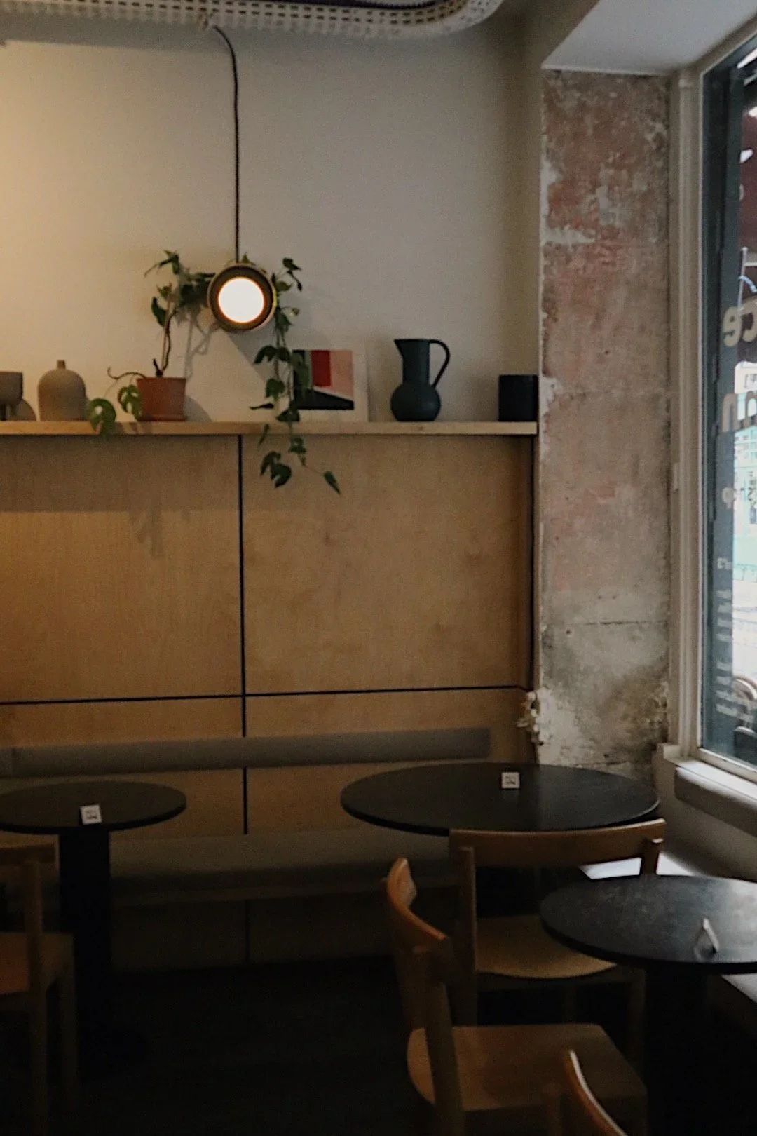 Intérieur d'un café avec des tables en bois rond, un mur en brique partiellement visible, une étagère avec des décorations, et une fenêtre laissant entrer de la lumière naturelle.