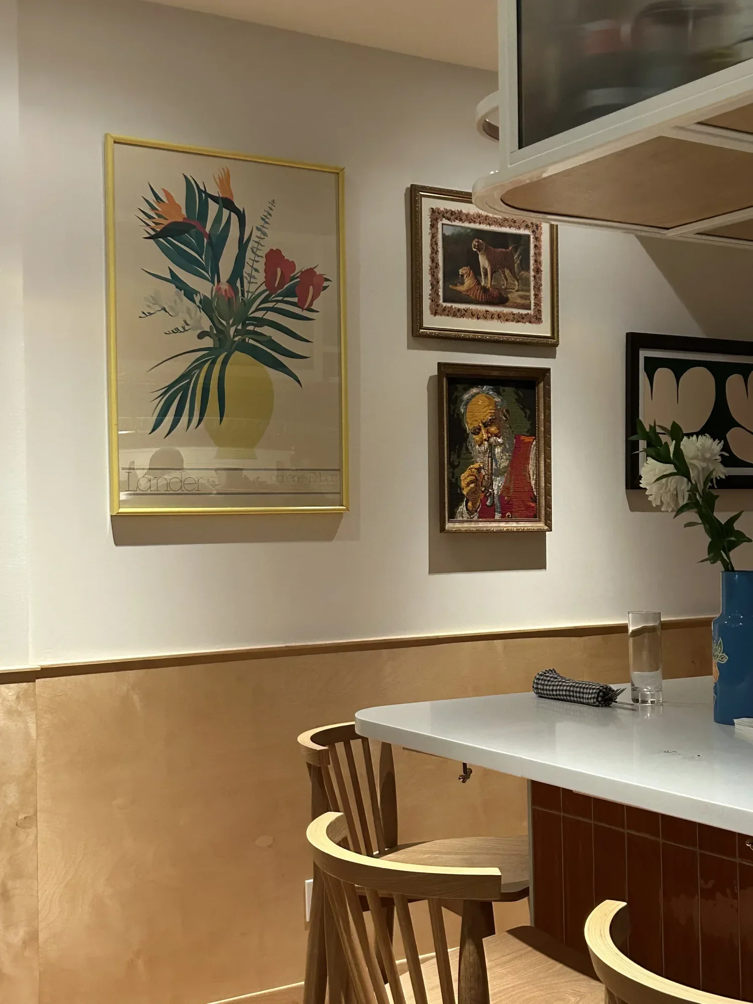 Une partie d'une salle à manger ou un coin salon avec un mur décoré de plusieurs tableaux, une table blanche avec un vase de fleurs et un verre d'eau, ainsi que deux chaises en bois.