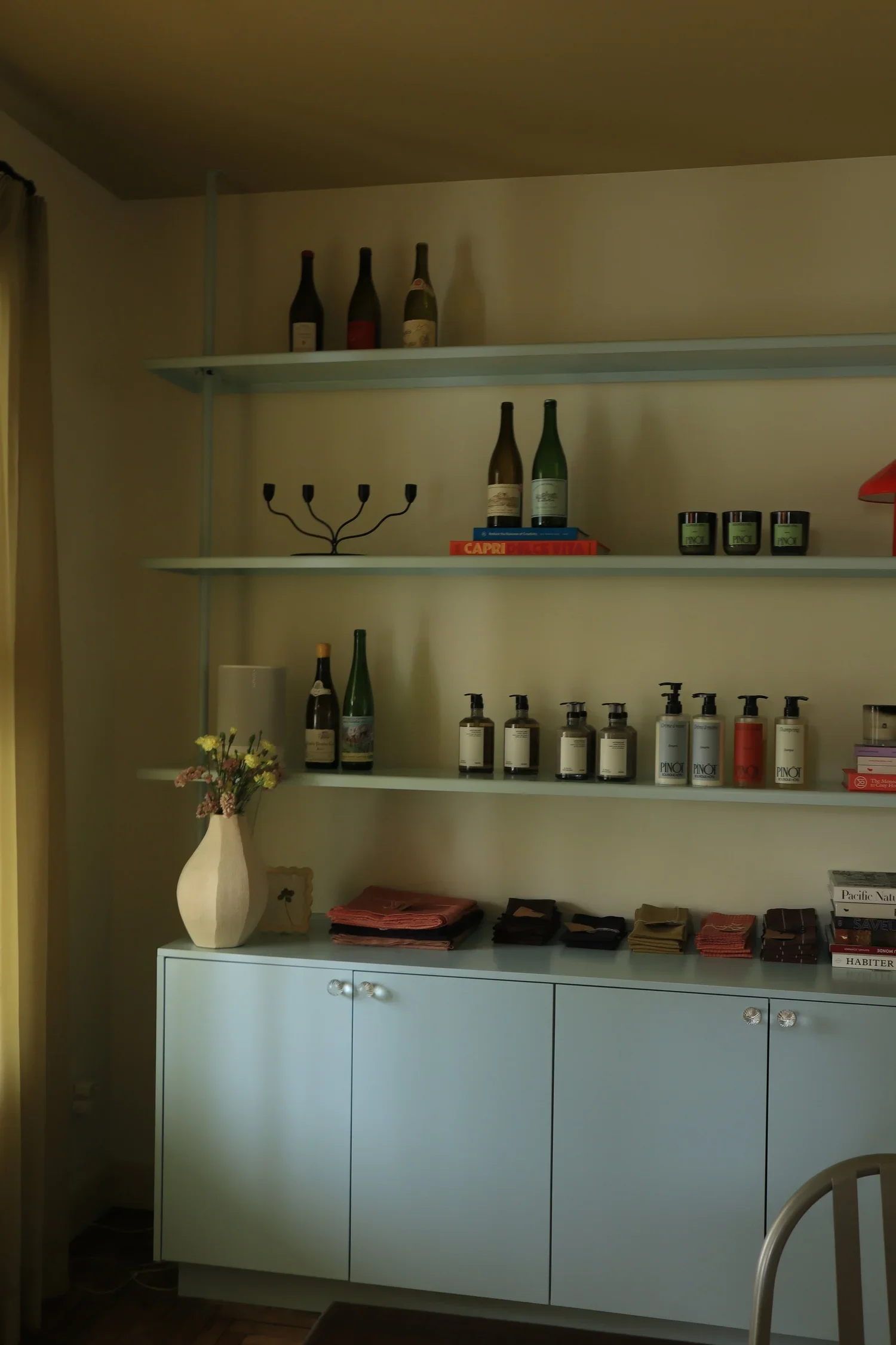 Une étagère blanche avec des bouteilles de vin, un chandelier, des pots de crème, des livres et un vase avec des fleurs, positionnée au-dessus d'un meuble blanc avec des livres et des tissus colorés. 