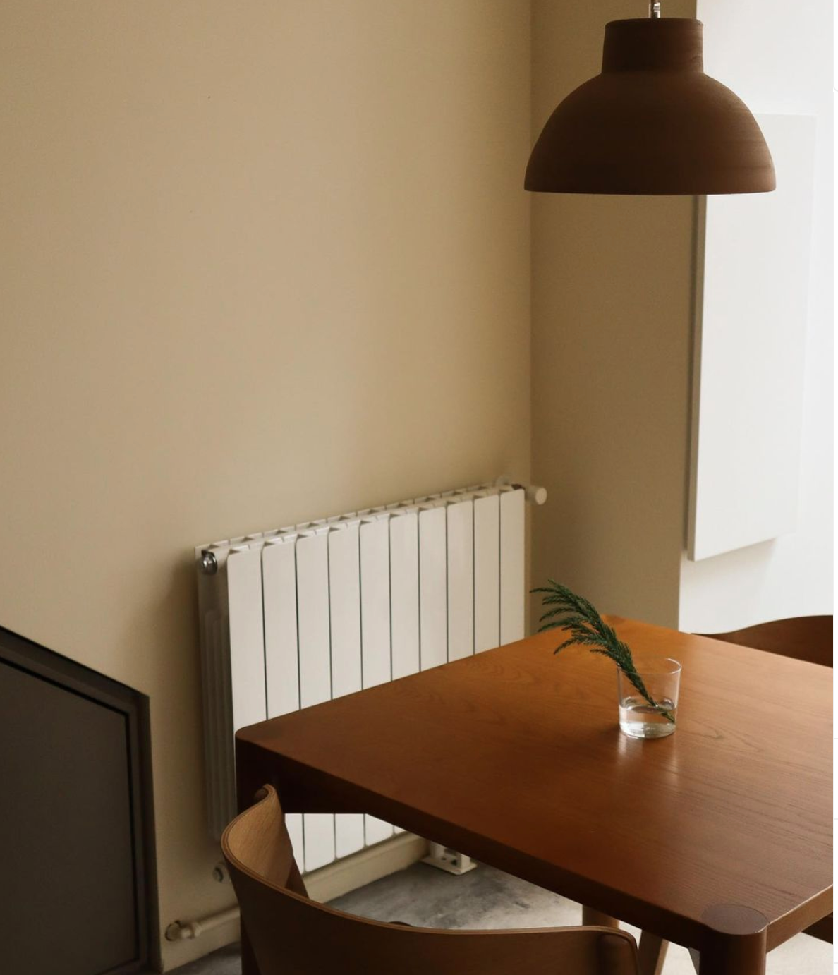 Table en bois avec un verre contenant une branche verte, suspension en bois au plafond, radiateur blanc, murs beige, meuble blanc