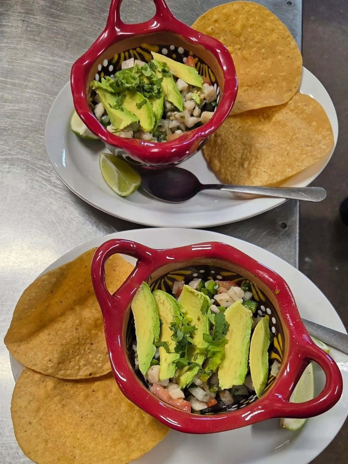 Two plates of Mexican food with tostadas, avocado slices, chopped raw onion and cilantro, lime wedges, and a small bowl of ceviche garnished with avocado and cilantro.