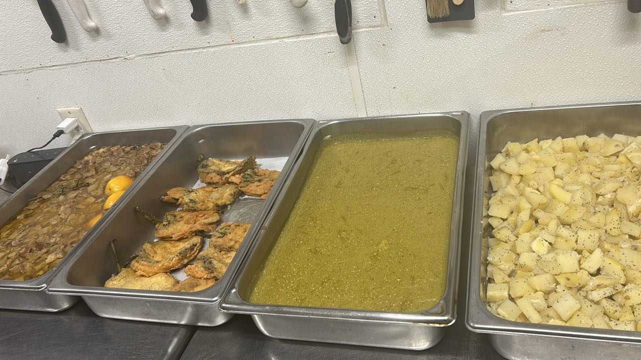 Buffet-style food trays with cooked meat, fried fish, green soup, and potato salad.