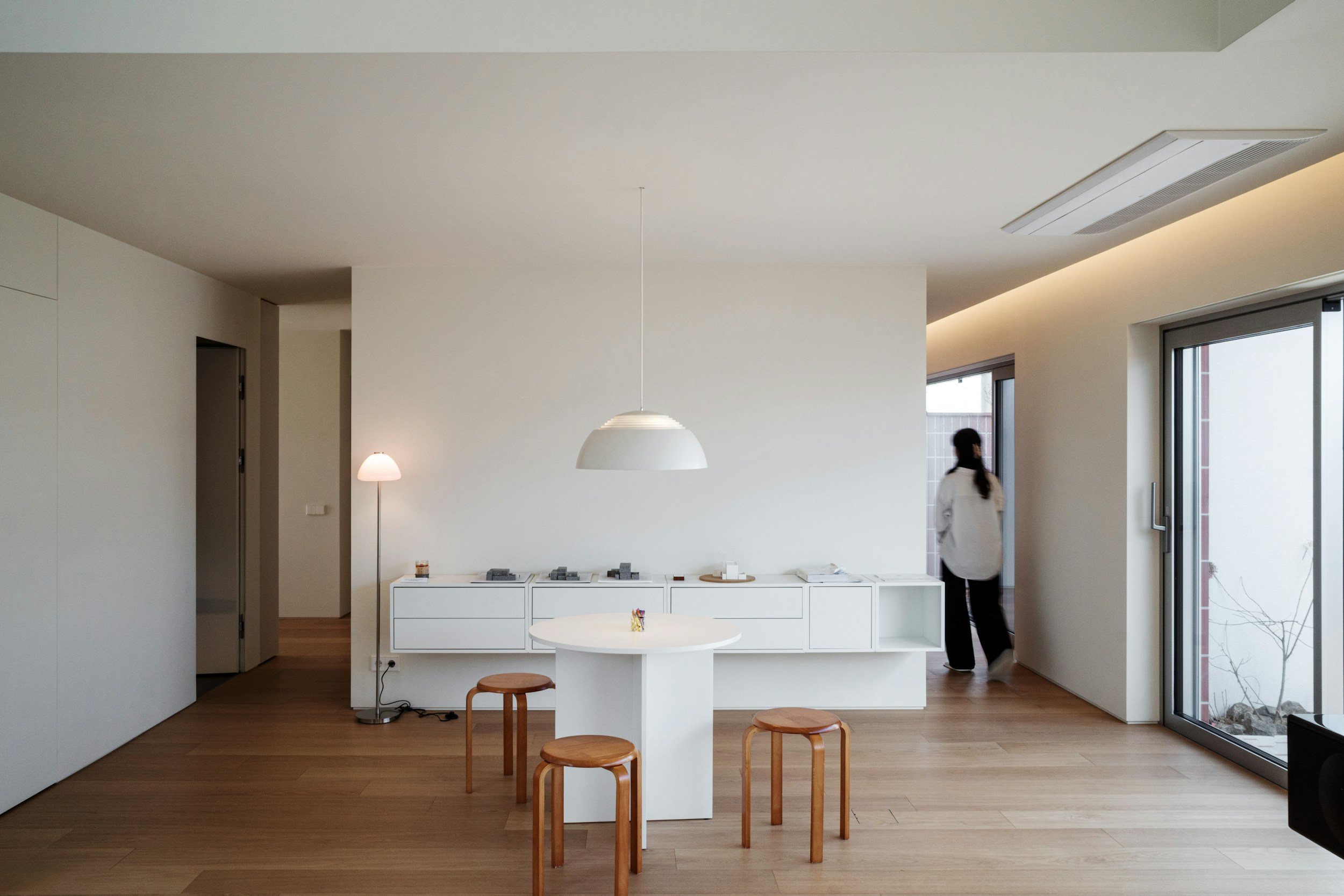 Minimalist kitchen with white cabinets and a small round table with three wooden stools, large sliding glass door, woman in white shirt walking outside, and wooden floor.