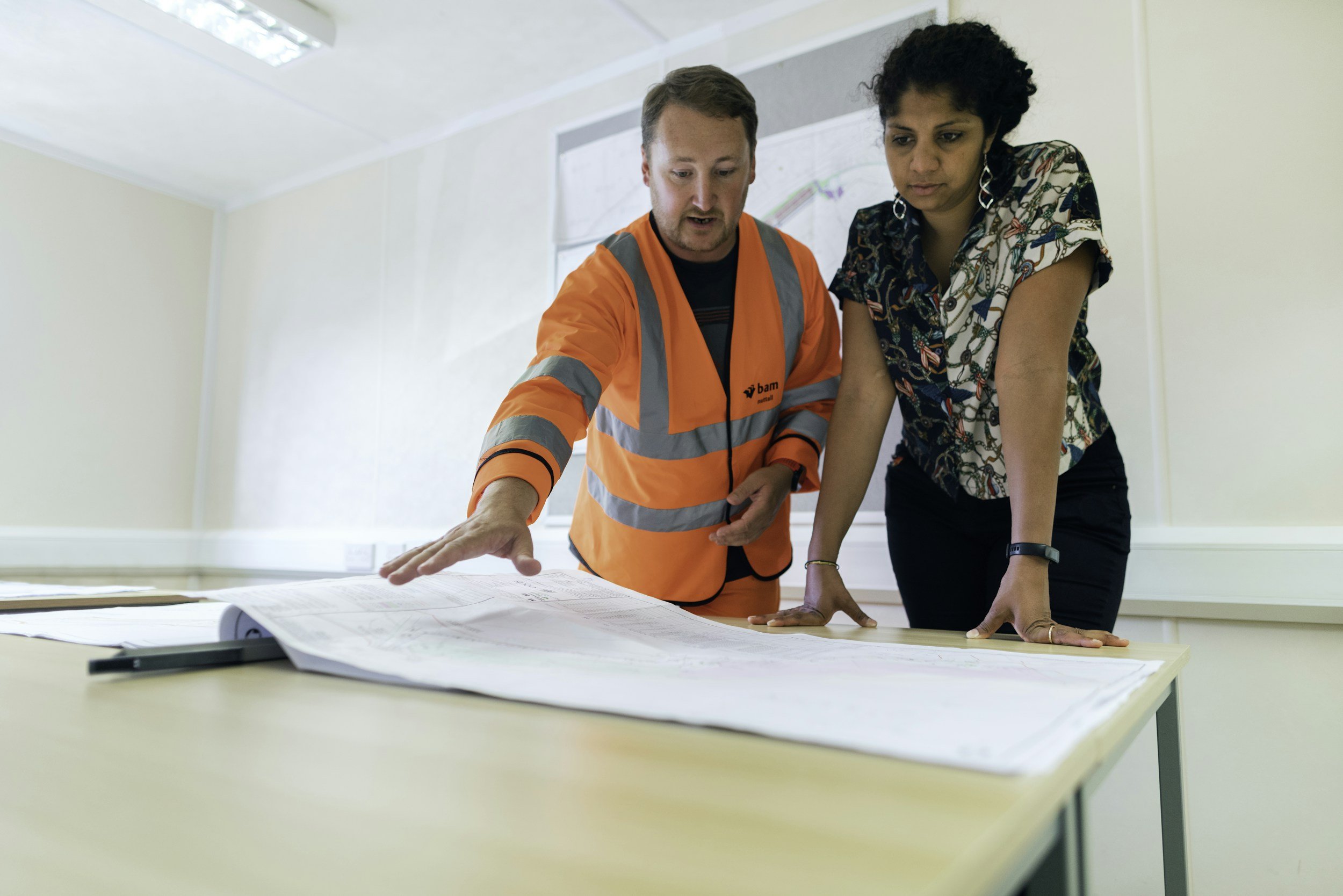 A man in an orange safety vest and a woman look at large plans or blueprints spread out on a table.