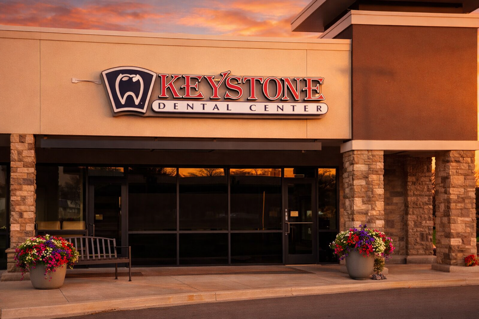 Exterior of Keystone Dental Center building with a sign, two large flower pots, and a bench during sunset.