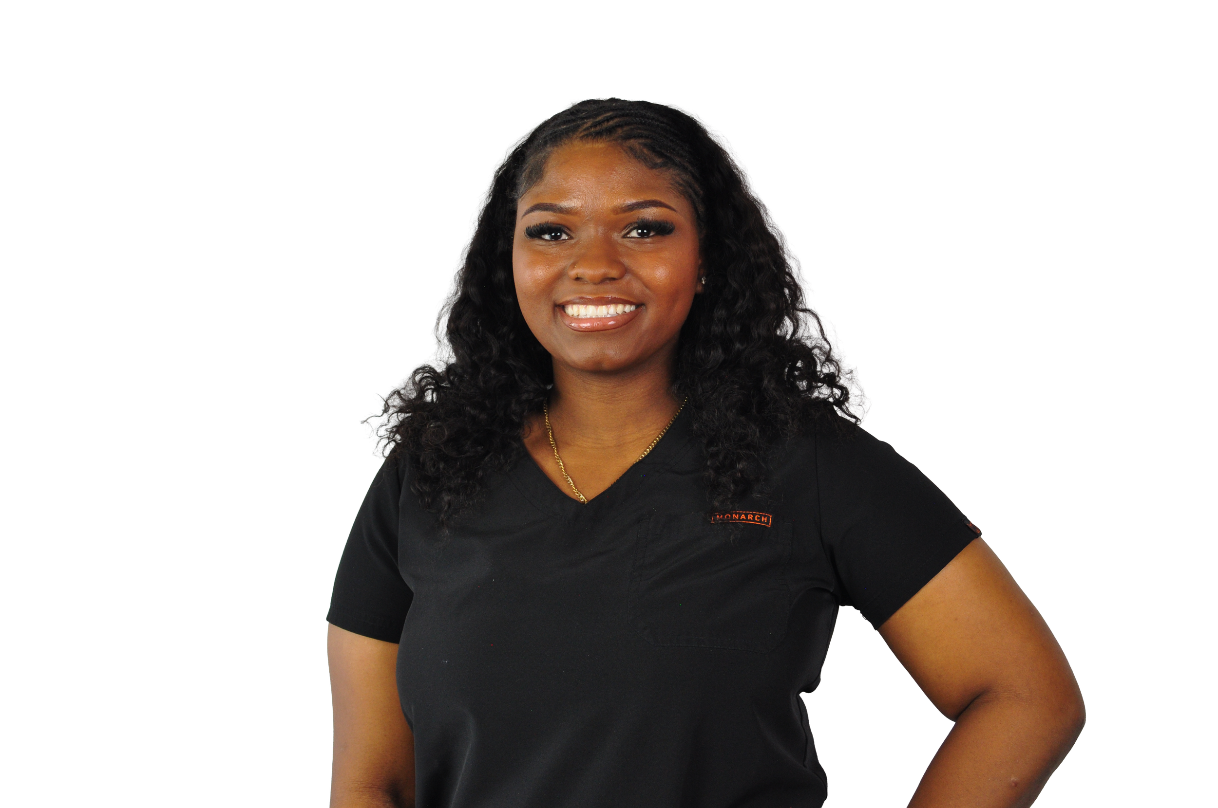 Smiling woman with dark curly hair wearing a black shirt and gold necklace, standing with hand on hip against a black background.