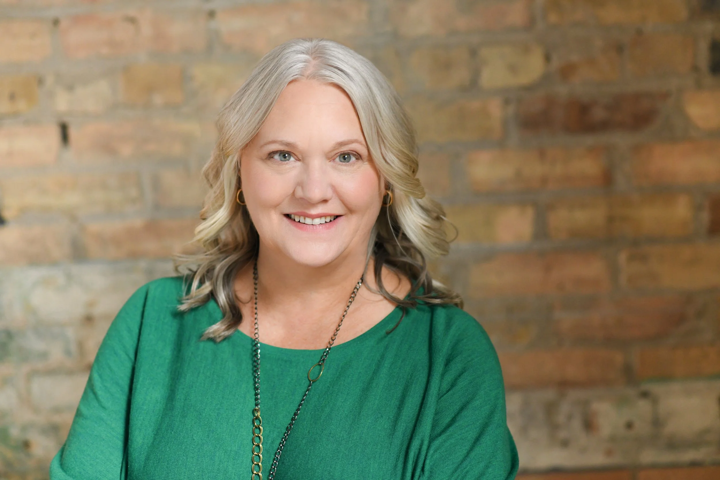 A smiling middle-aged woman with gray hair styled in loose waves, wearing a green top and a long necklace, standing in front of a brick wall.