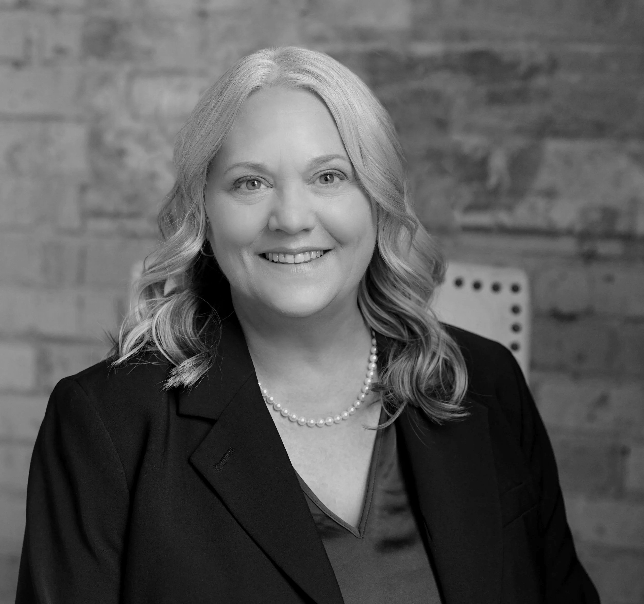 Black and white portrait of a woman with blonde hair styled in loose waves, wearing a pearl necklace and a dark blazer, smiling against a blurred indoor background.