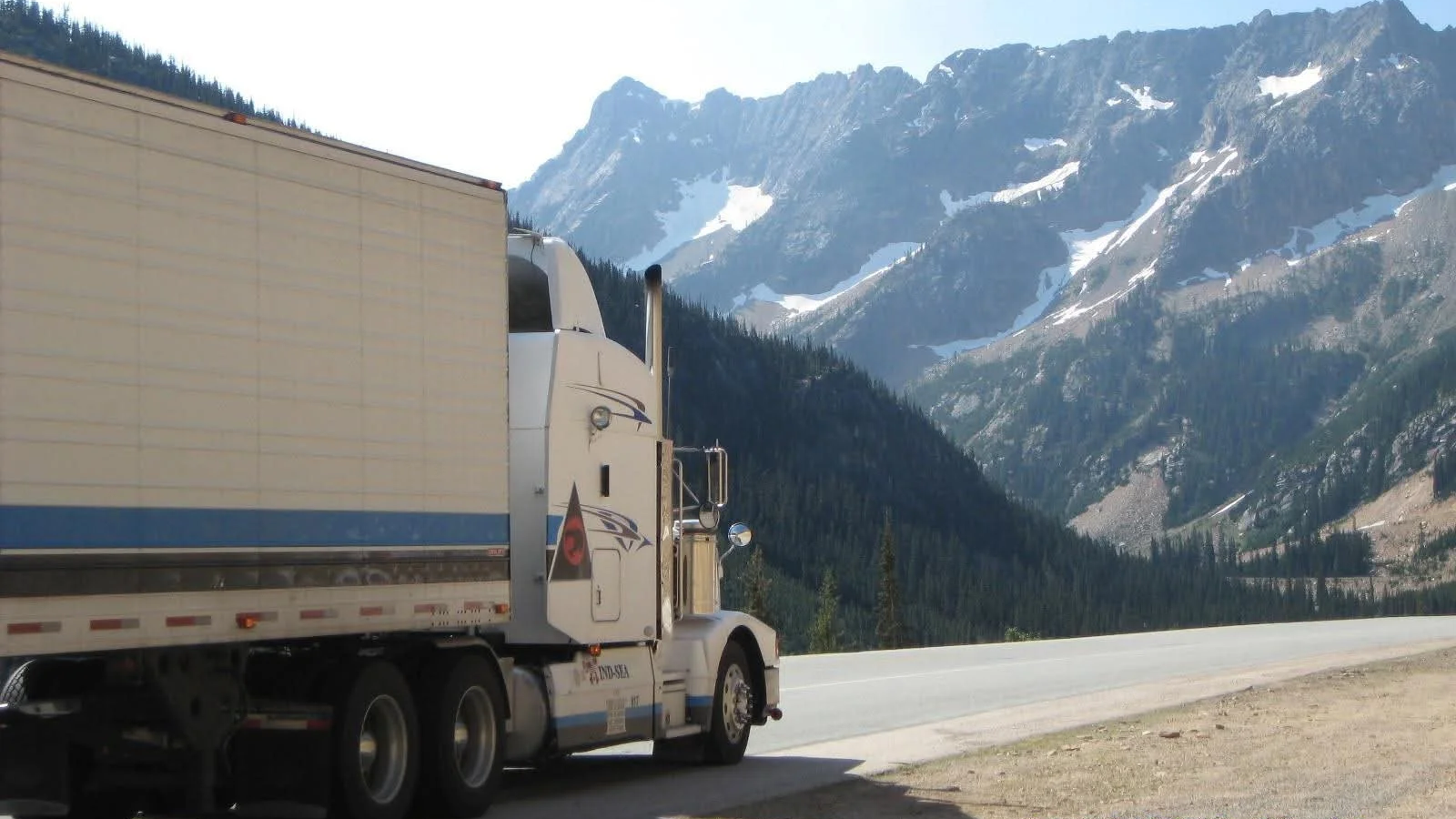 A semi-truck parked on the side of a mountain road with snow-capped mountains and forested slopes in the background.
