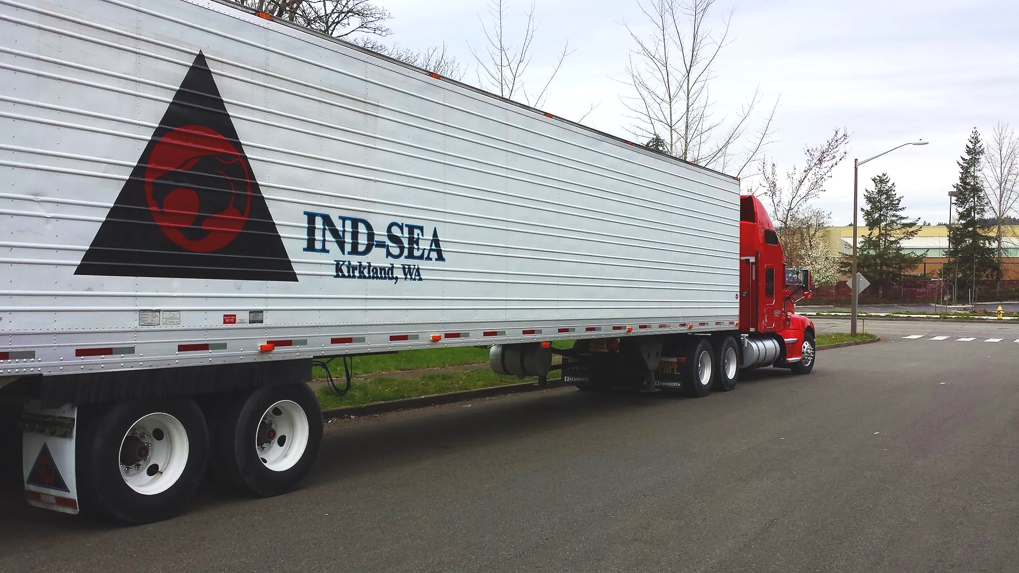 A semi-truck with a red cab and white trailer parked on a street near trees and buildings in Kirkland, Washington.