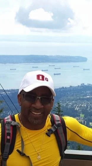 Man wearing a white cap, sunglasses, and a yellow shirt smiling outdoors with a scenic view of water and ships in the background.
