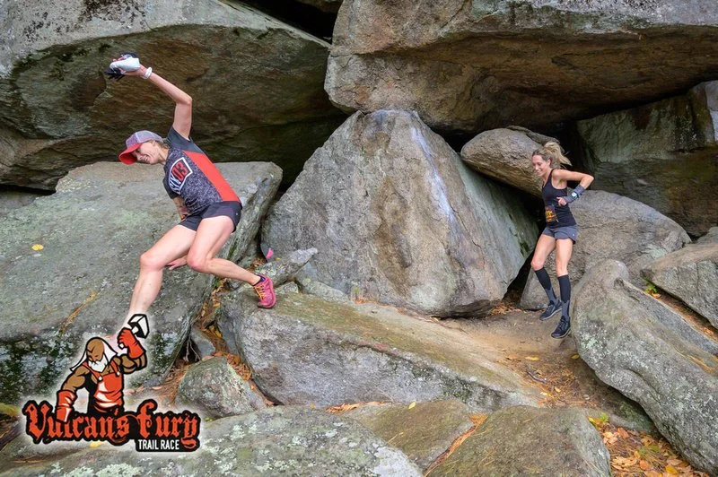 A person in athletic gear climbing over large rocks in a trail race, with another person observing nearby, in a rugged outdoor setting.