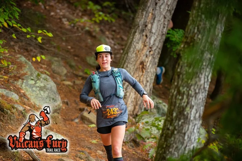 A woman trail running through a wooded forest, wearing a grey long sleeve shirt, black shorts, a yellow and white cap, and a backpack, with a race bib number 243, during a Vulcan Fury Trail Race.