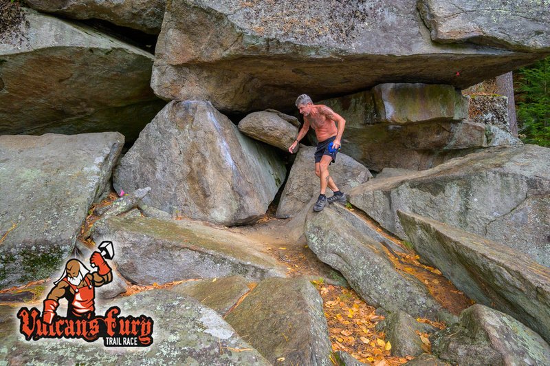 A shirtless man in shorts and hiking boots walking on a large rock under a massive rock formation in a forested area.