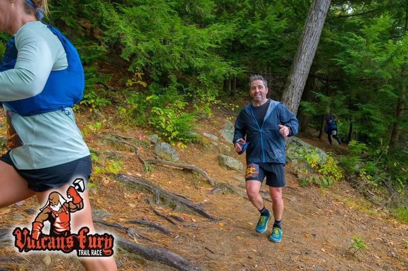 A man running on a forest trail during a trail race, with another runner partially visible on the left and a third runner in the background, surrounded by green trees.