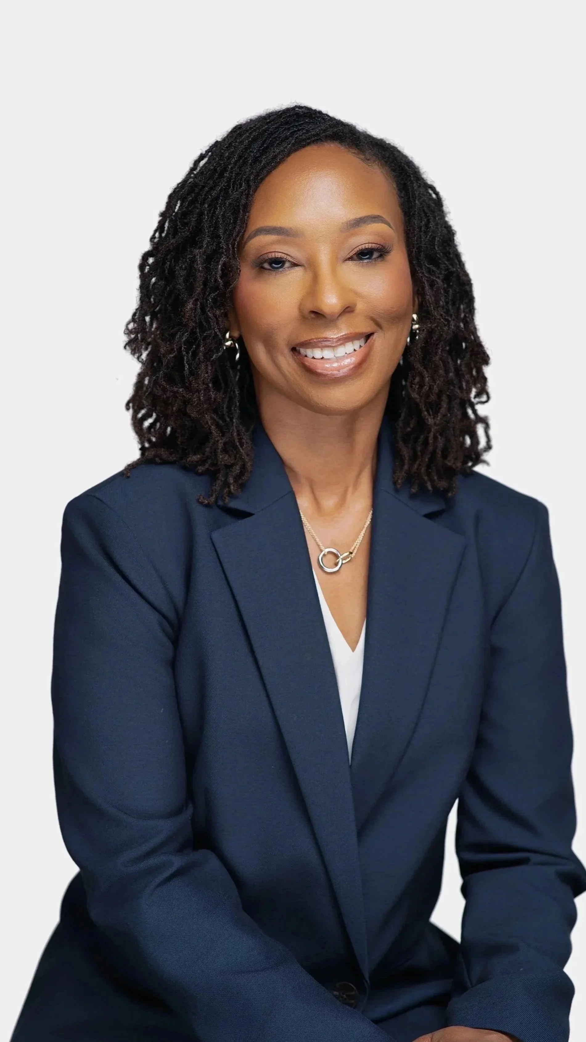 A professional woman with dark, curly shoulder-length hair, wearing a navy blue blazer, a white top, and jewelry, smiling in front of a plain light gray background.