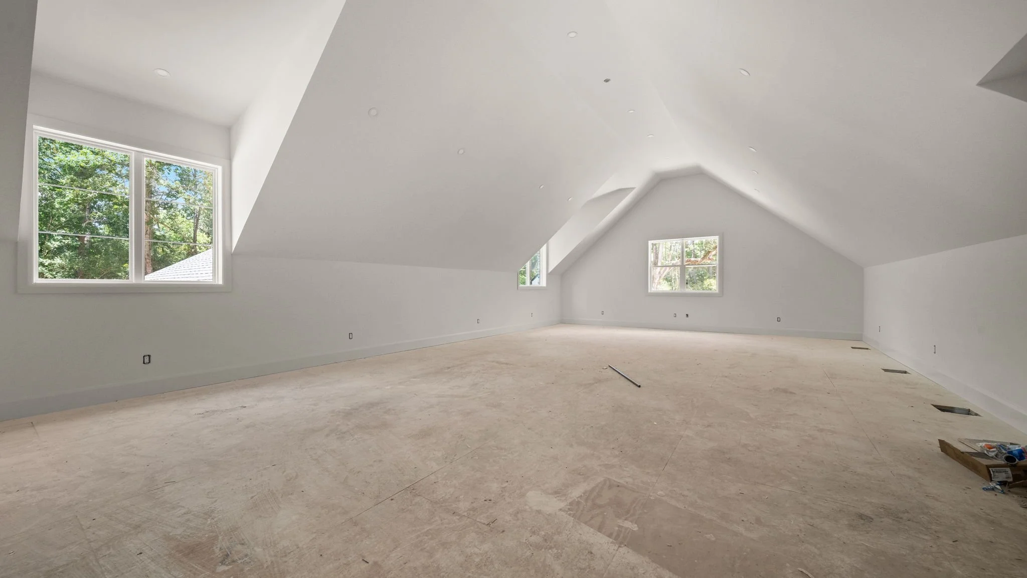 An empty attic or loft space with white walls and ceiling, three windows letting in natural light, and a bare unfinished floor with some construction tools.
