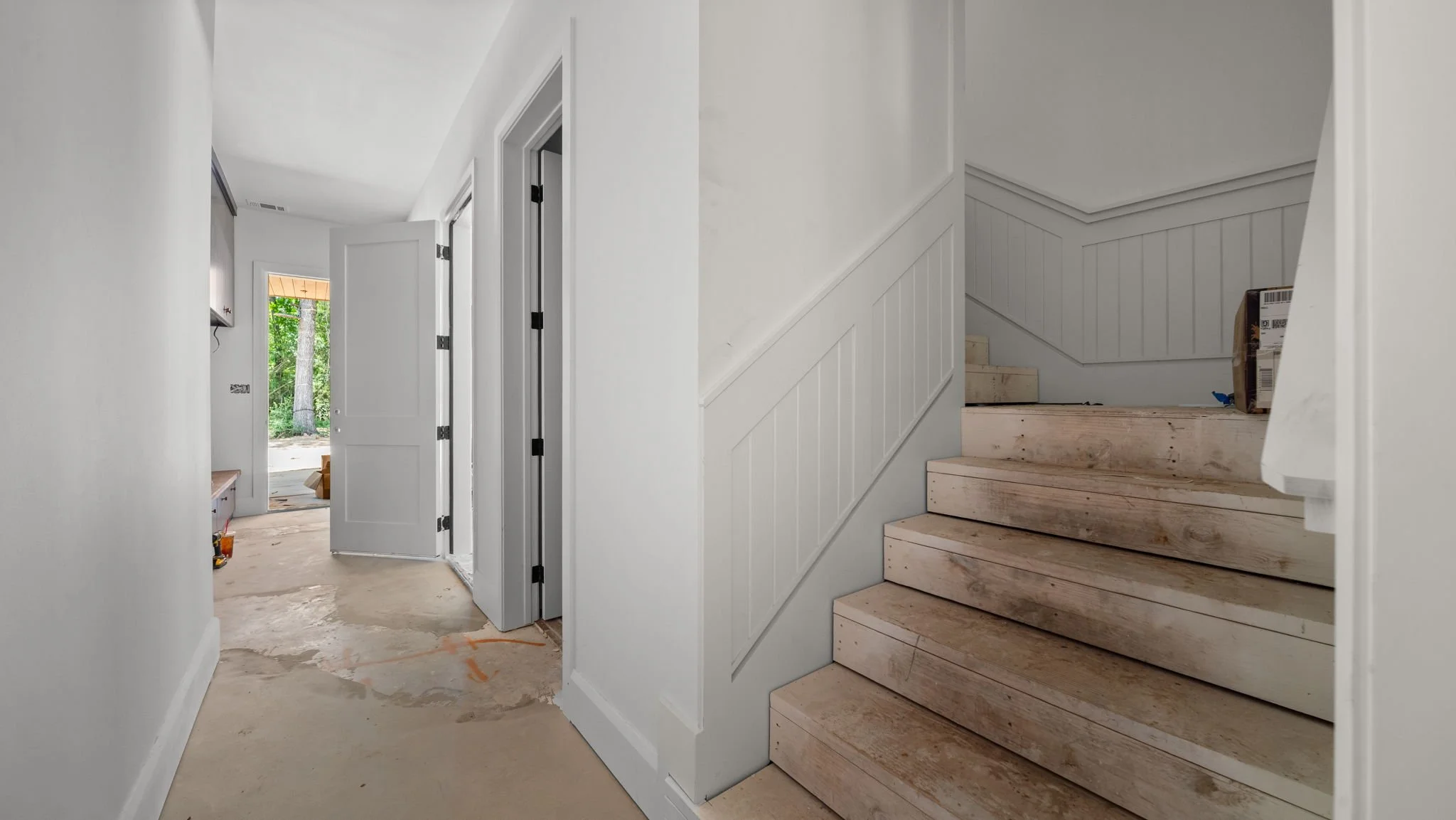 Interior view of a house staircase with unfinished stairs and white walls, with a hallway leading to a door and window showing greenery outside.