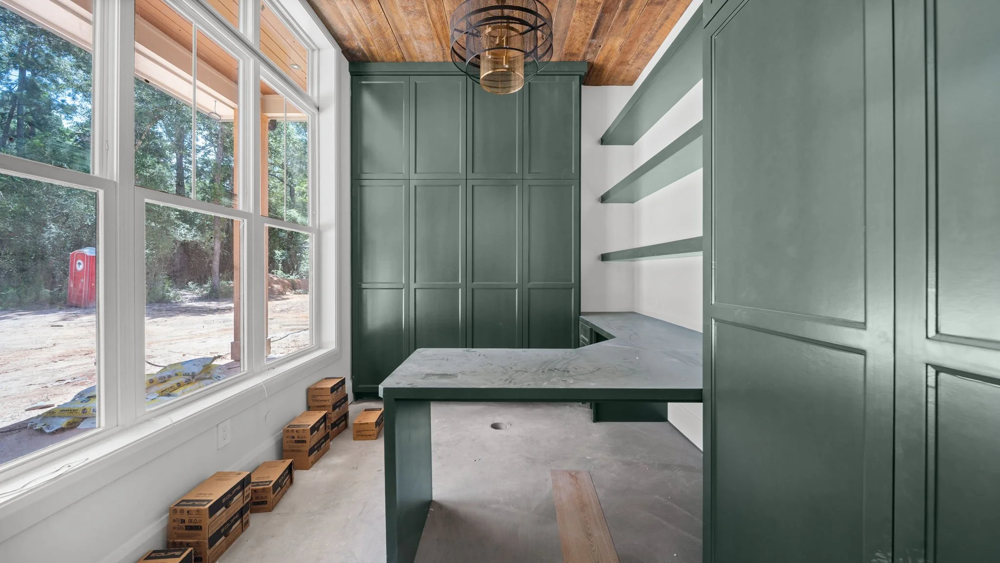 Interior of a kitchen with green cabinetry, white countertops, large window showing outdoor trees, wooden ceiling, and some boxes on the floor.