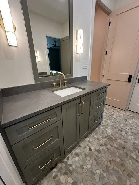 Bathroom vanity with grey cabinets, gold handles, a grey countertop, and a white sink beneath a large mirror with wall-mounted light fixtures.