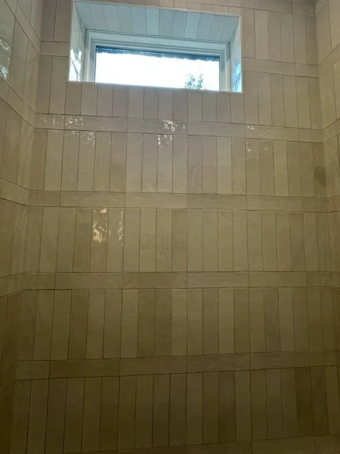 Close-up of a tiled shower wall with rectangular beige tiles and a small window at the top.