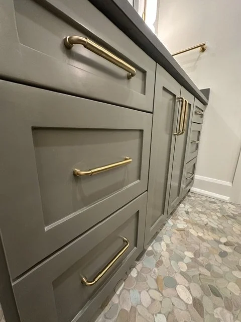 Close-up view of gray bathroom vanity cabinet with gold handles and patterned tile floor.