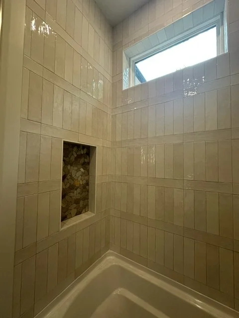 Shower wall with beige tiles and a built-in niche, with a window above.