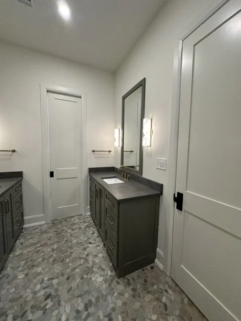 Bathroom with a single sink vanity, dark countertop, large mirror, light fixtures, white walls, and patterned tile floor.