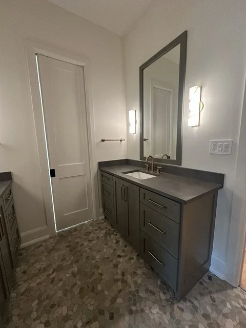Bathroom vanity with a grey cabinet, a white sink, a large mirror, and two wall-mounted lights, with a door on the left and a patterned tile floor.