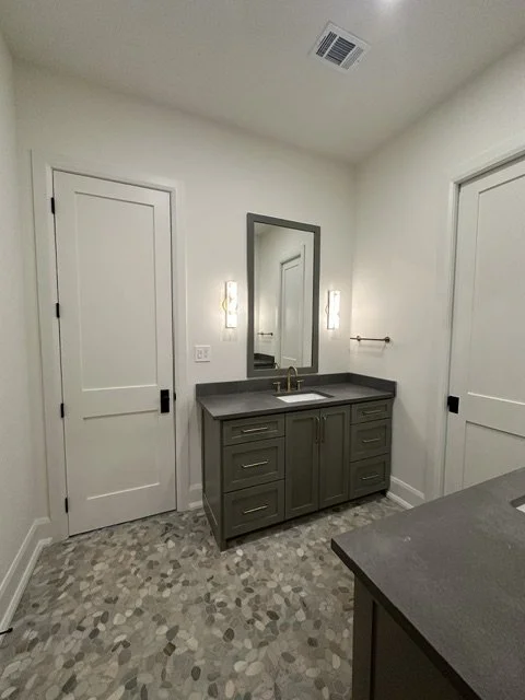 Bathroom with gray vanity, mirror, two wall-mounted light fixtures, two white doors, and a patterned tile floor.