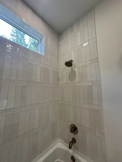 Bathroom shower with beige tiled walls, a small window, a rain showerhead, and a faucet at the bottom.