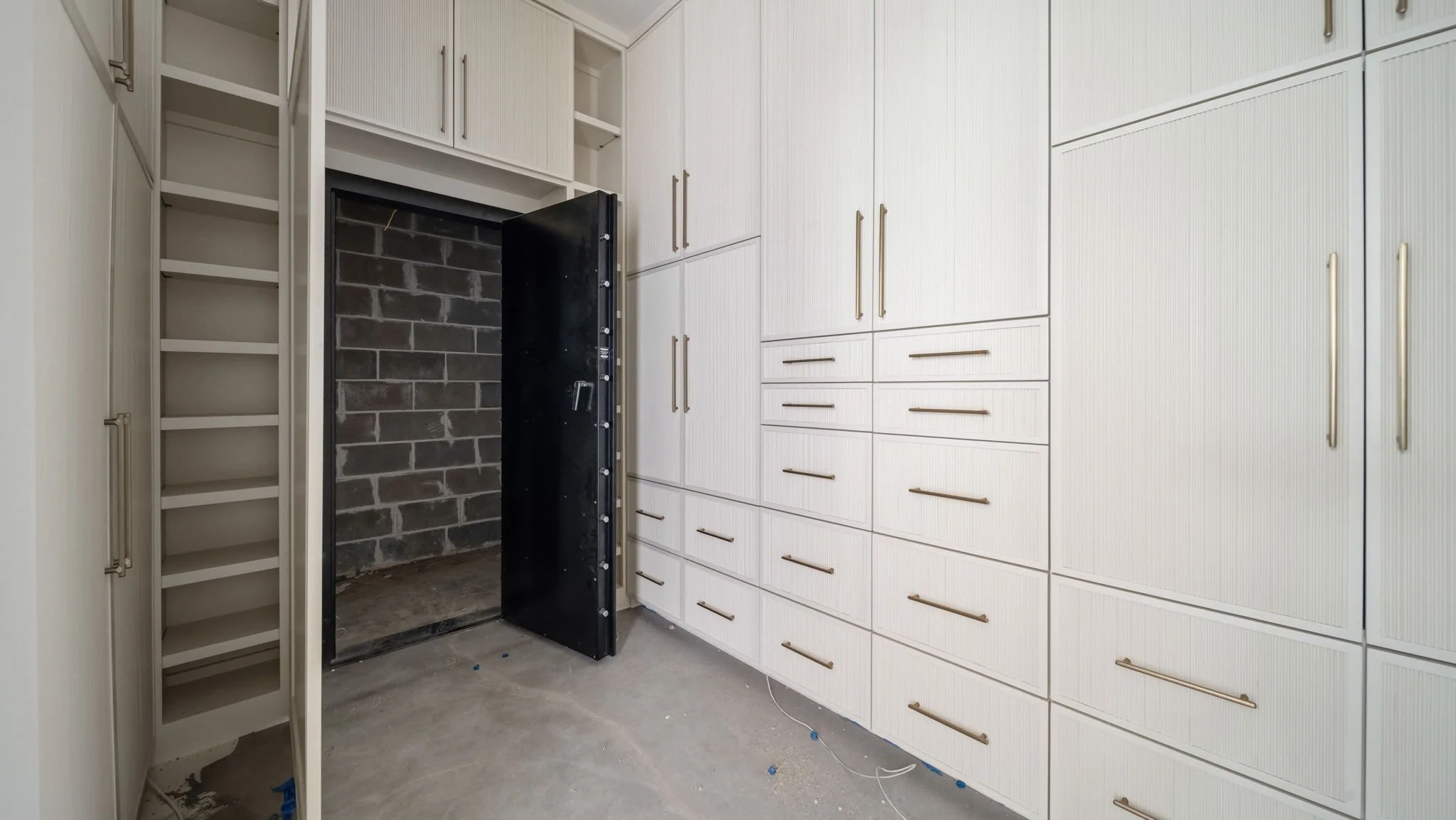A walk-in closet with white cabinets, gold handles, an open safe, and a concrete floor.