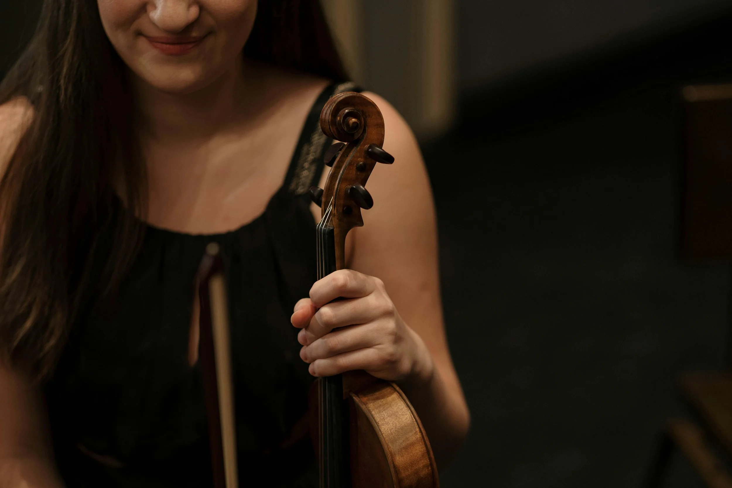 A woman holding a violin and smiling with a dark background.
