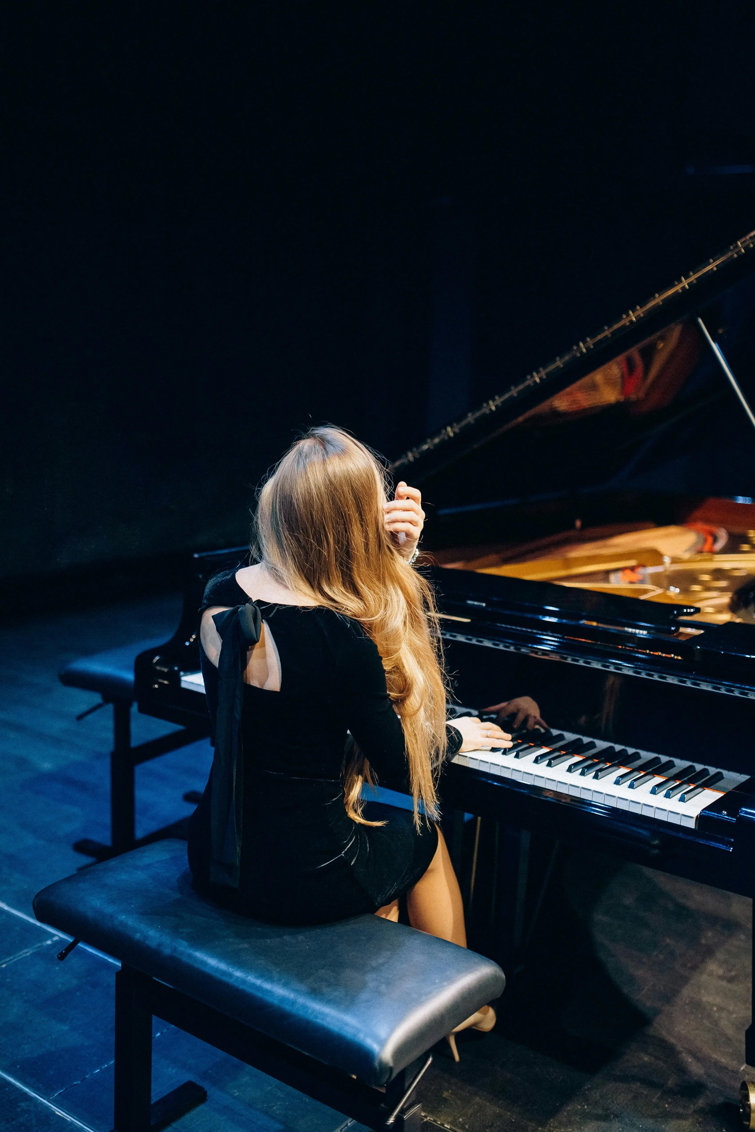 A woman with long blonde hair in a black dress sitting at a grand piano on stage, covering her face with her hand, with a black background.