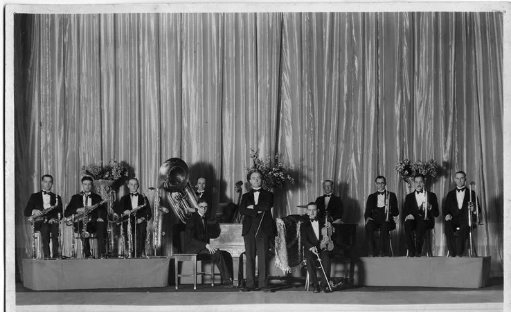 A black and white photo of a jazz band performing on stage with a curtain backdrop. The band members are dressed in tuxedos and are playing various instruments including saxophones, a tuba, and a drum set. Some members are standing, while others are seated, with flower arrangements placed on either side of the stage.