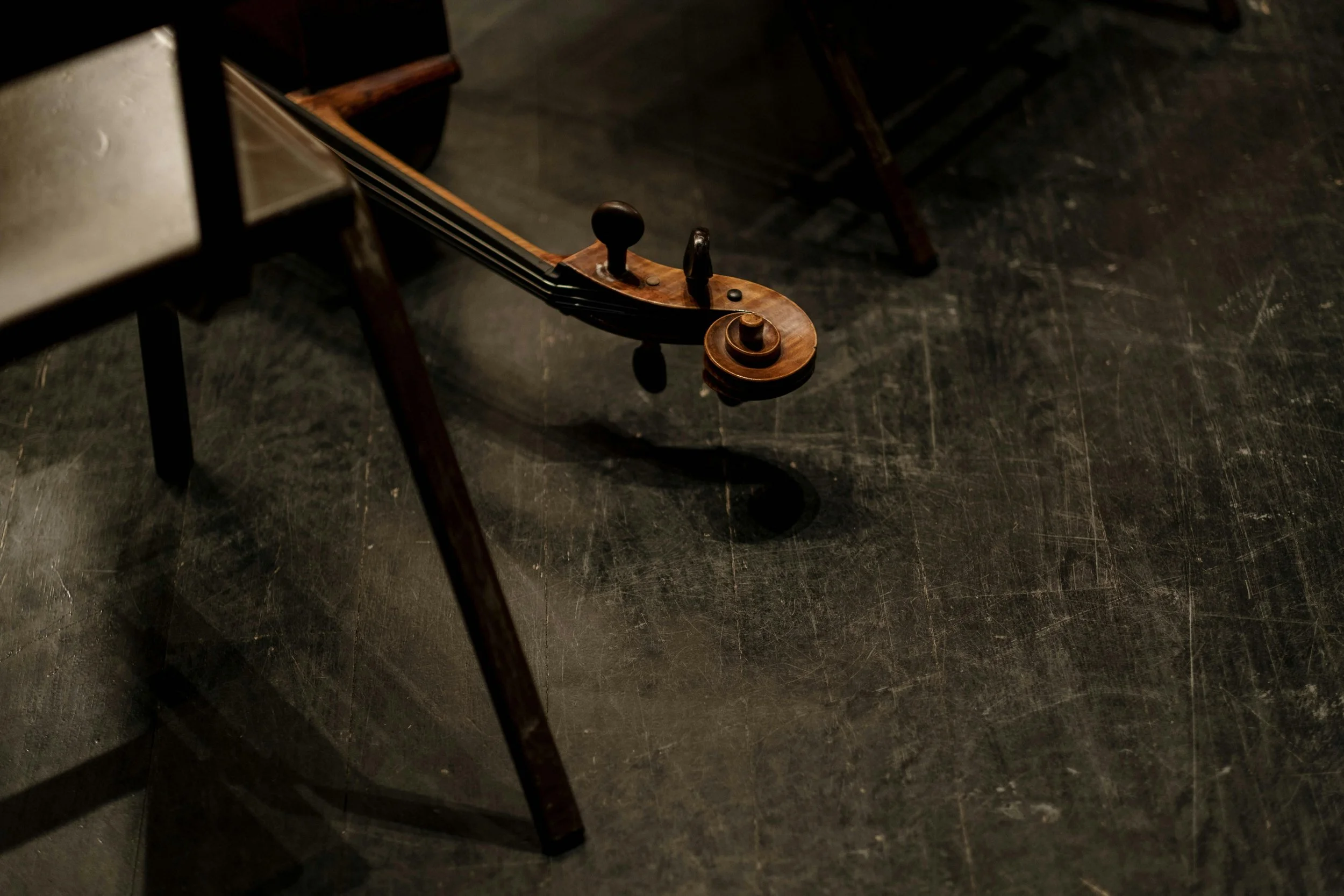 A wooden cello sits on a dark, worn stage floor, with its shadow cast underneath.