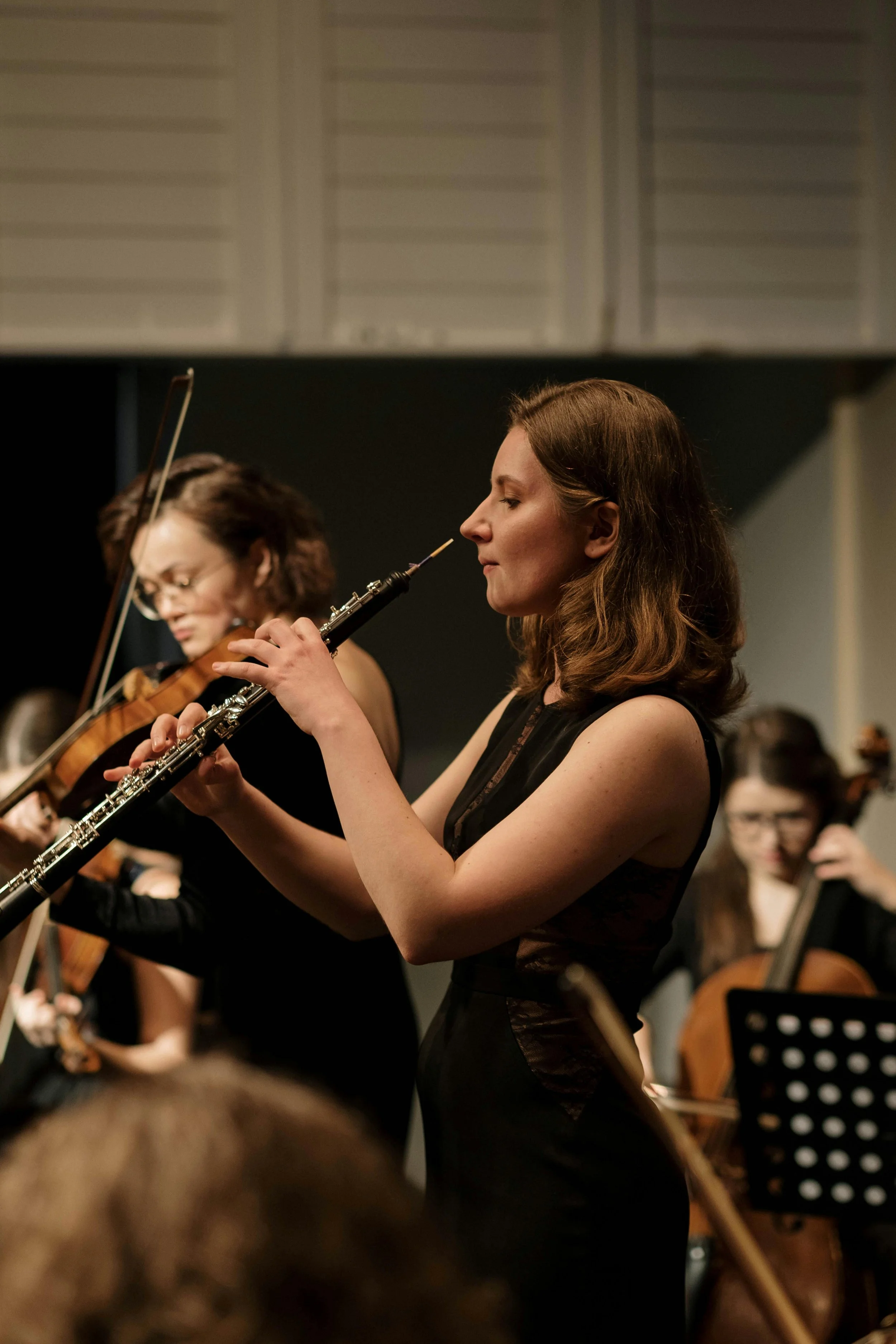 Women playing classical instruments in an orchestra, centered on a woman playing the flute, with other musicians in the background.