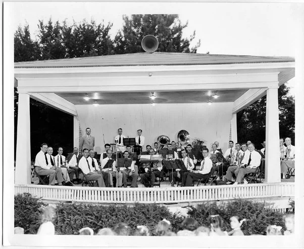 A black and white photo of a large outdoor band performing on a stage under a pavilion.