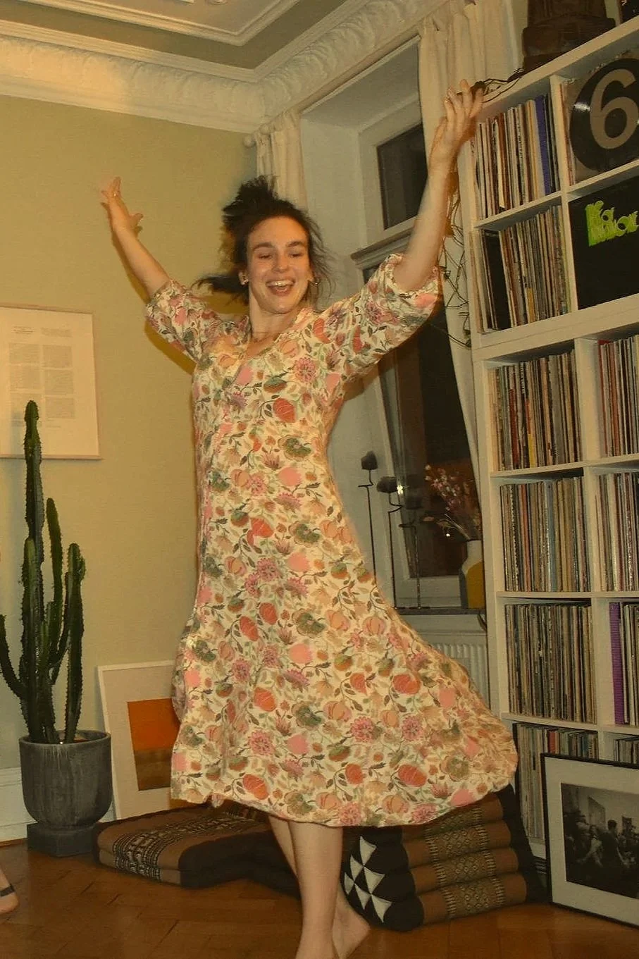 Eine lachende Frau in einem floral gemusterten Kleid tanzt in einem Wohnzimmer.