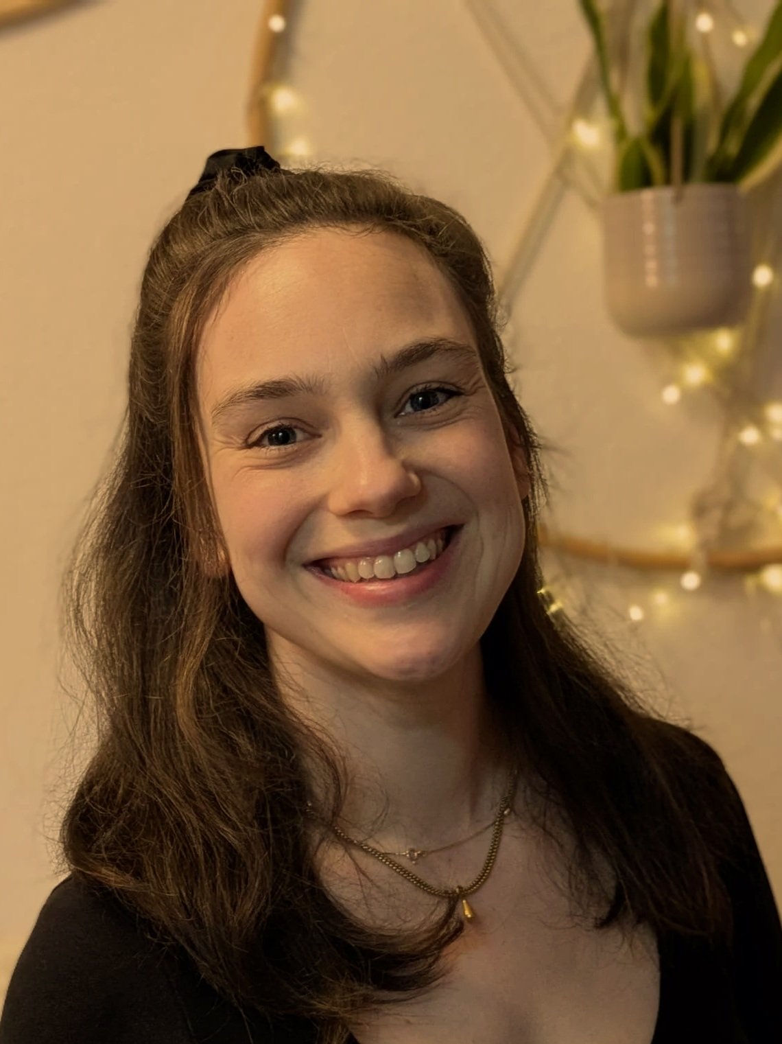 Lächelnde junge Frau mit braunen Haaren, schwarzen Oberteil, Goldkette und Krawattenart Frisur, im Hintergrund eine Pflanze und Dekoration mit Lichtern.