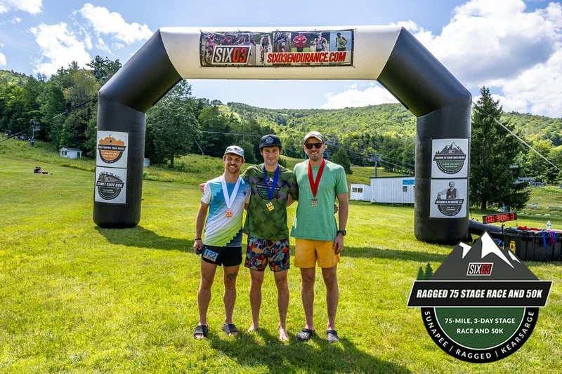 Three men with medals around their necks standing on grass in front of an inflatable archway at an outdoor race event, with green hills and some buildings in the background.