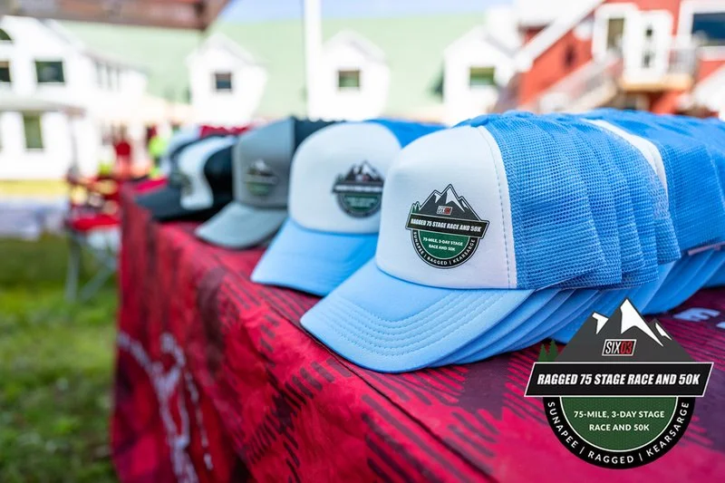 Multiple white and blue trucks hats with a logo for the 'Ragged 75 Stage Race and 50K' event on a red tablecloth in an outdoor setting, with houses and event tents in the background.