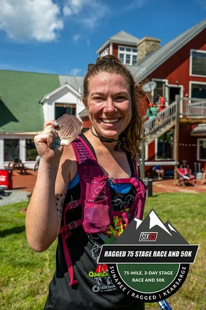 Woman smiling and holding a medal after a race, standing outdoors in front of colorful houses.